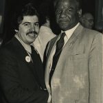 Two men stand side by side, one Latinx with a moustache, the other African American in a tan suit.