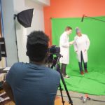 2 teens stand in front of a green screen, another teen holds a microphone on a long arm and a fourth teen takes video